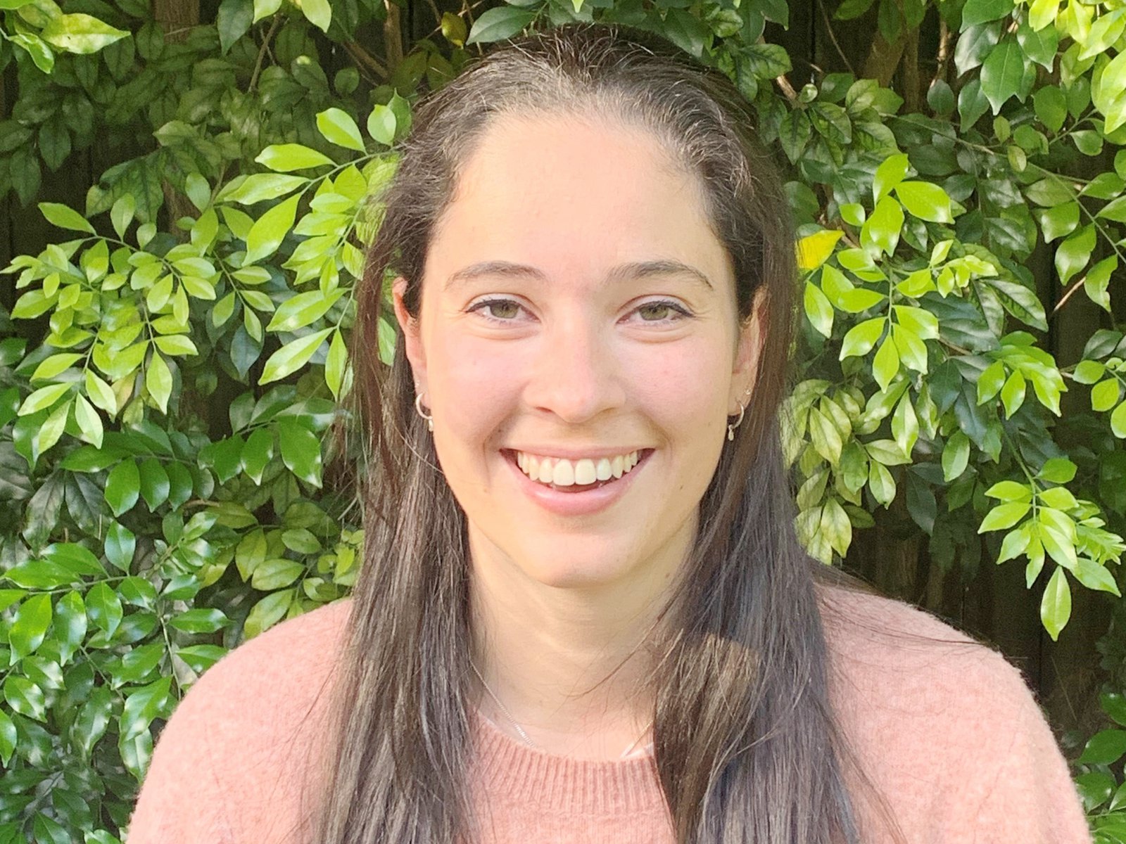 A woman with long dark hair smiles at the camera wearing a pink top against a leafy green background