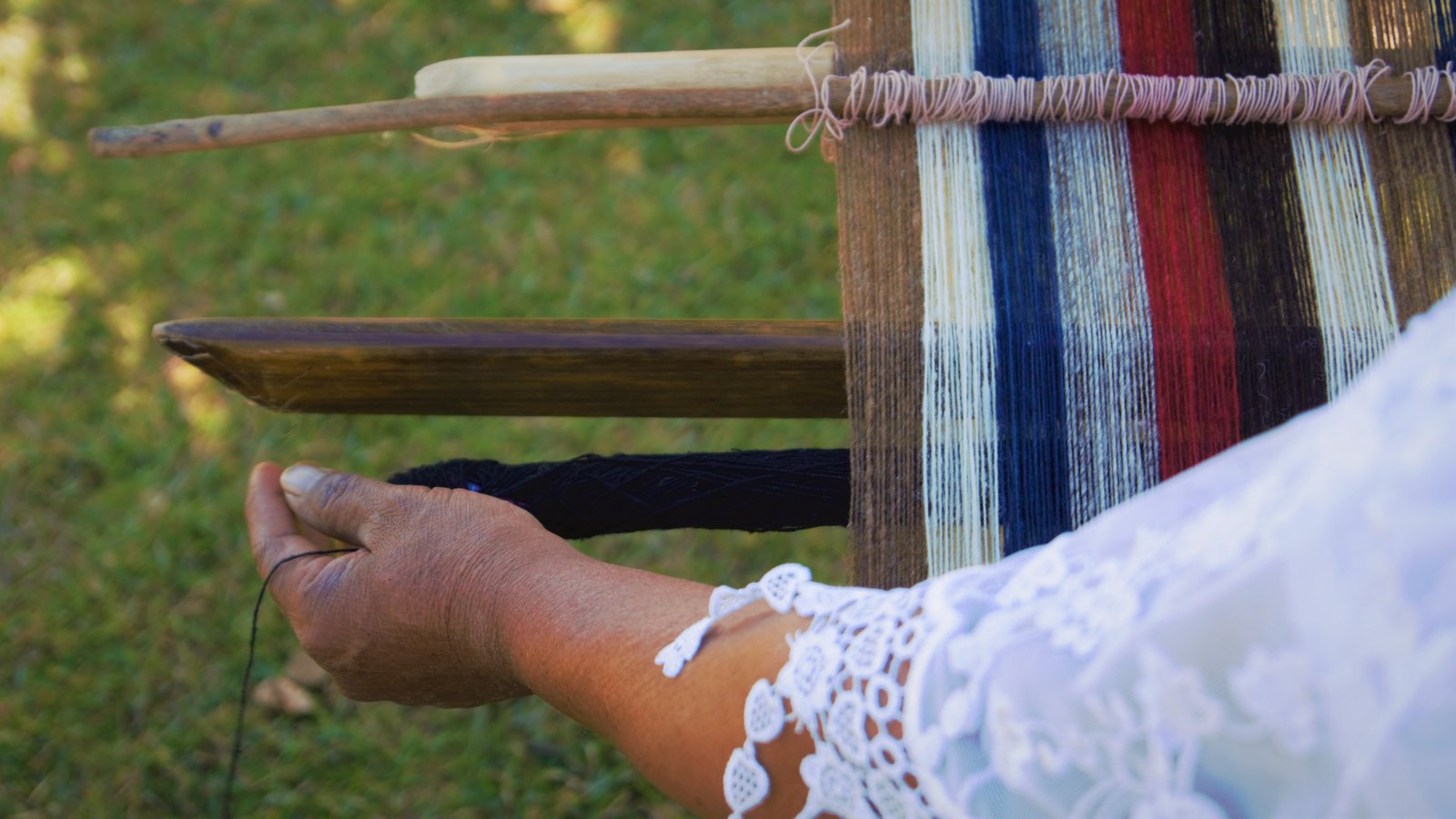 Close-up of hands weaving dark yarn through a traditional wooden wool loom with multicoloured thread