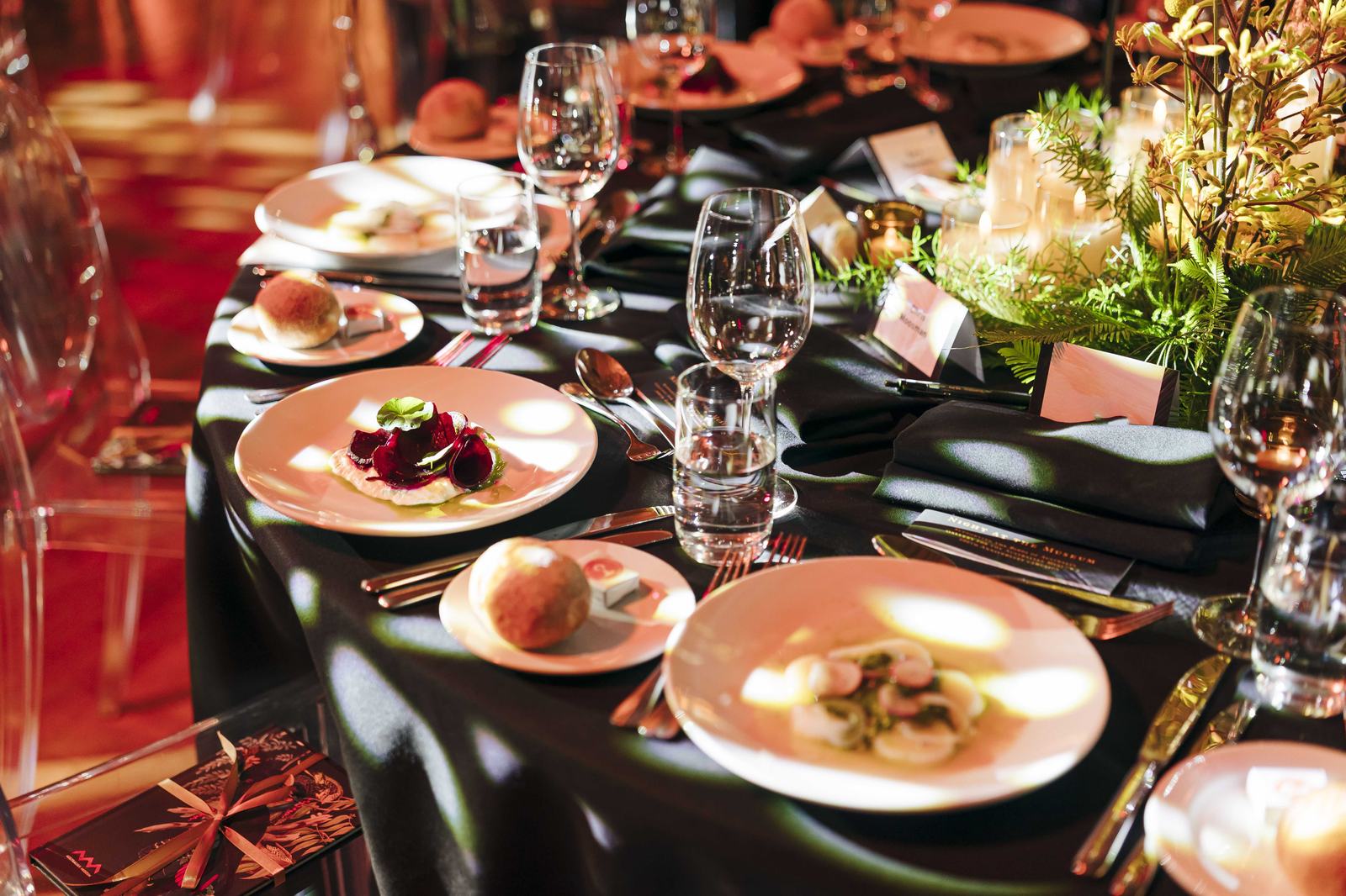 Long banquet table set with white plates, clear glasses, silverware, and a green floral centerpiece.