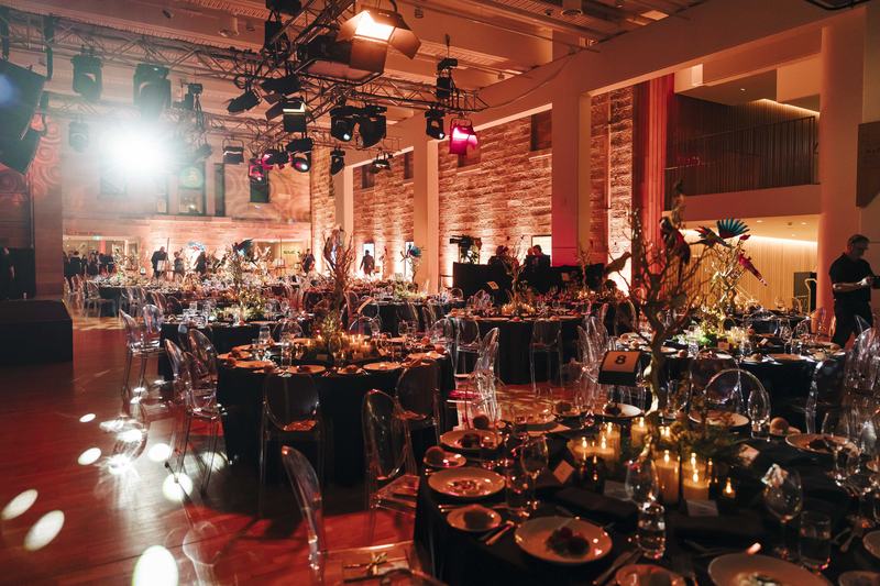 Large hall filled with round tables, each covered in black cloth and decorated with plates, candles, and various decorations.