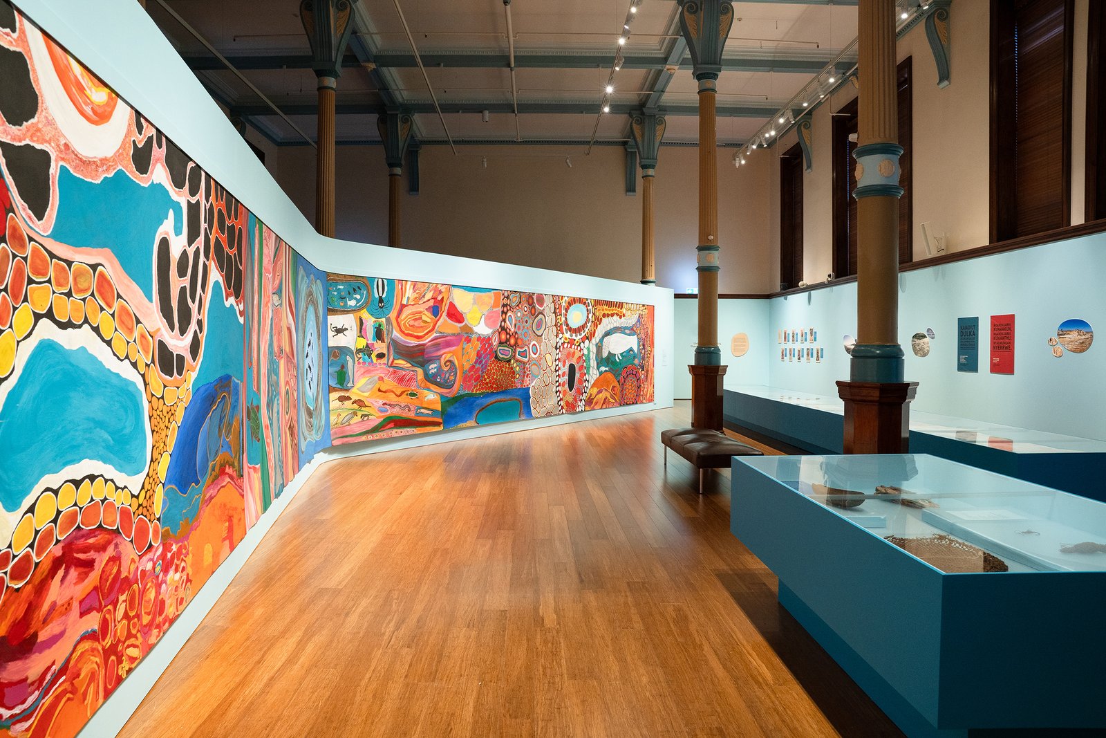 Gallery view of First Nations exhibition with mural artwork, wooden columns and cultural objects.