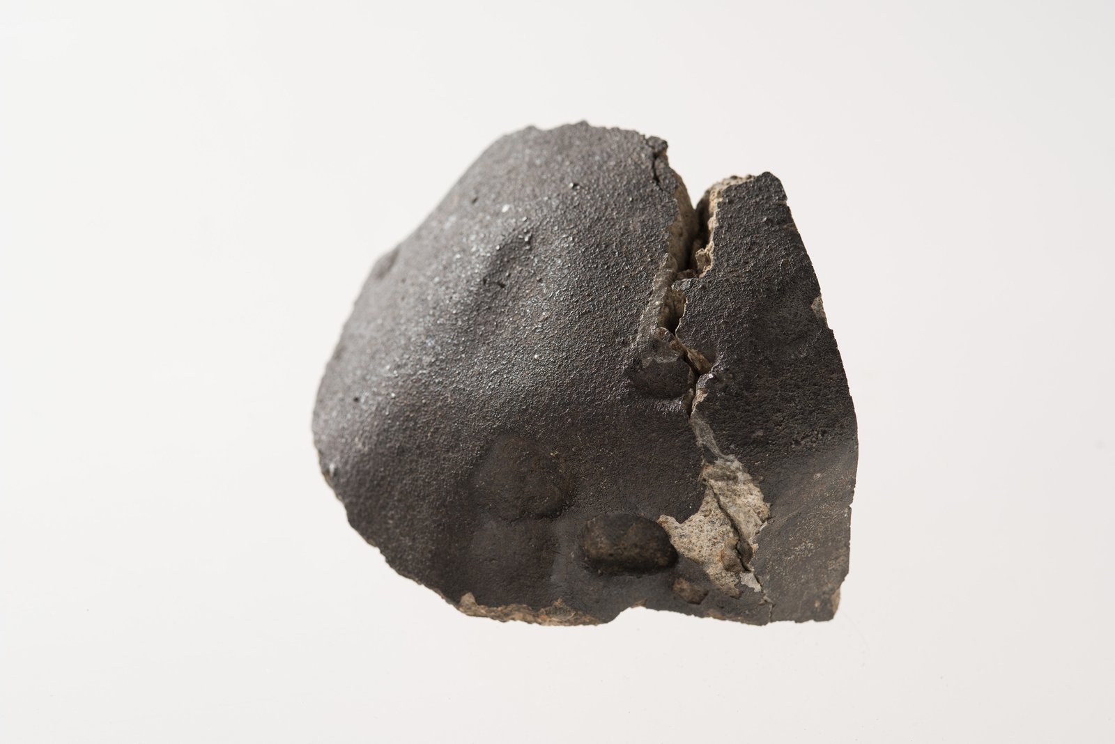 A dark grey irregular meteorite with a smooth fusion crust and fractured surface.