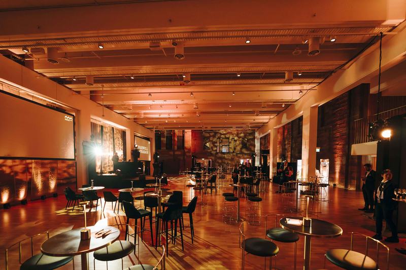 Large museum hall with wooden floors filled with bar tables and chairs.