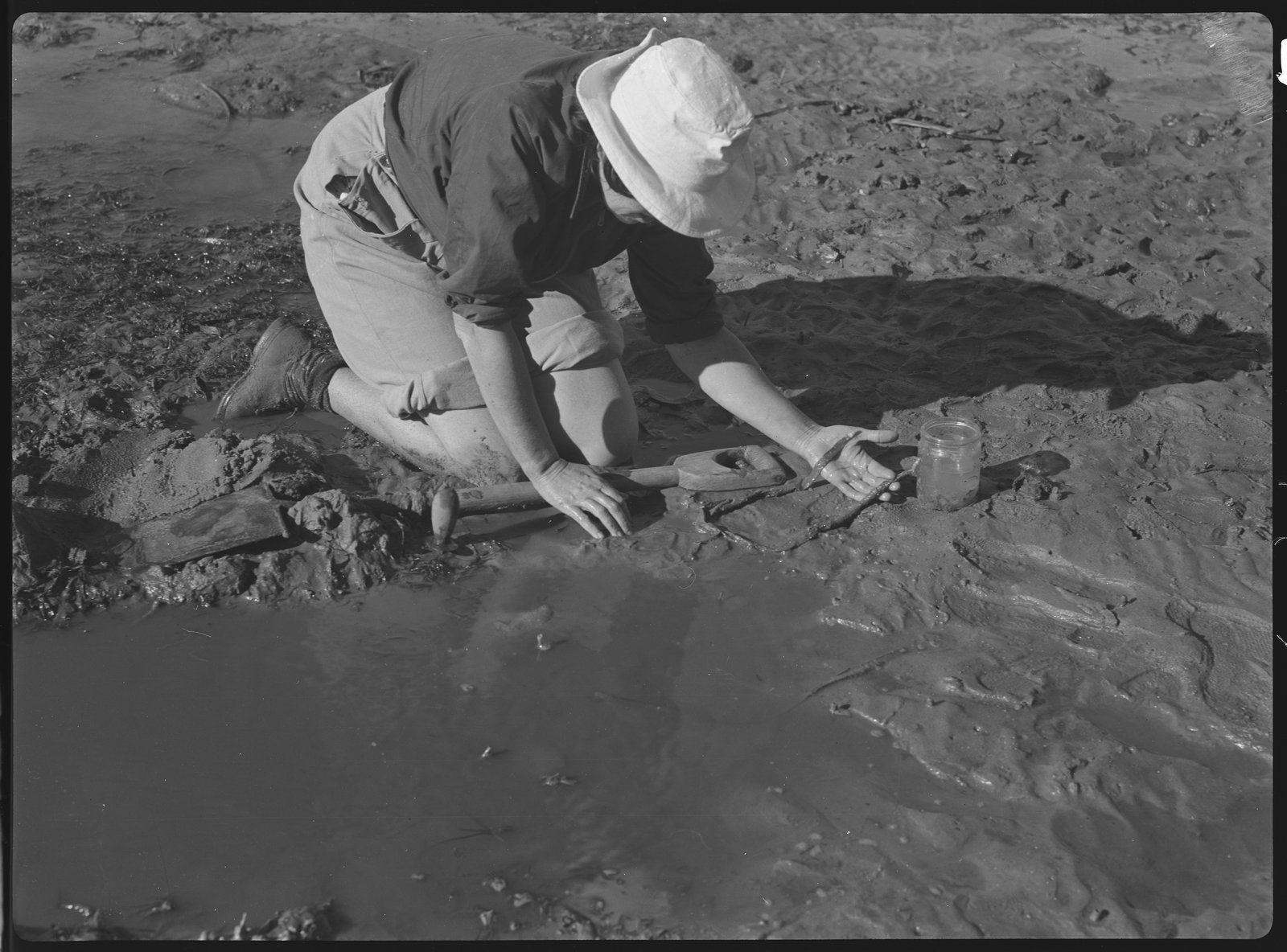 Black and white photograph of a woman crouching in sand with equipment.