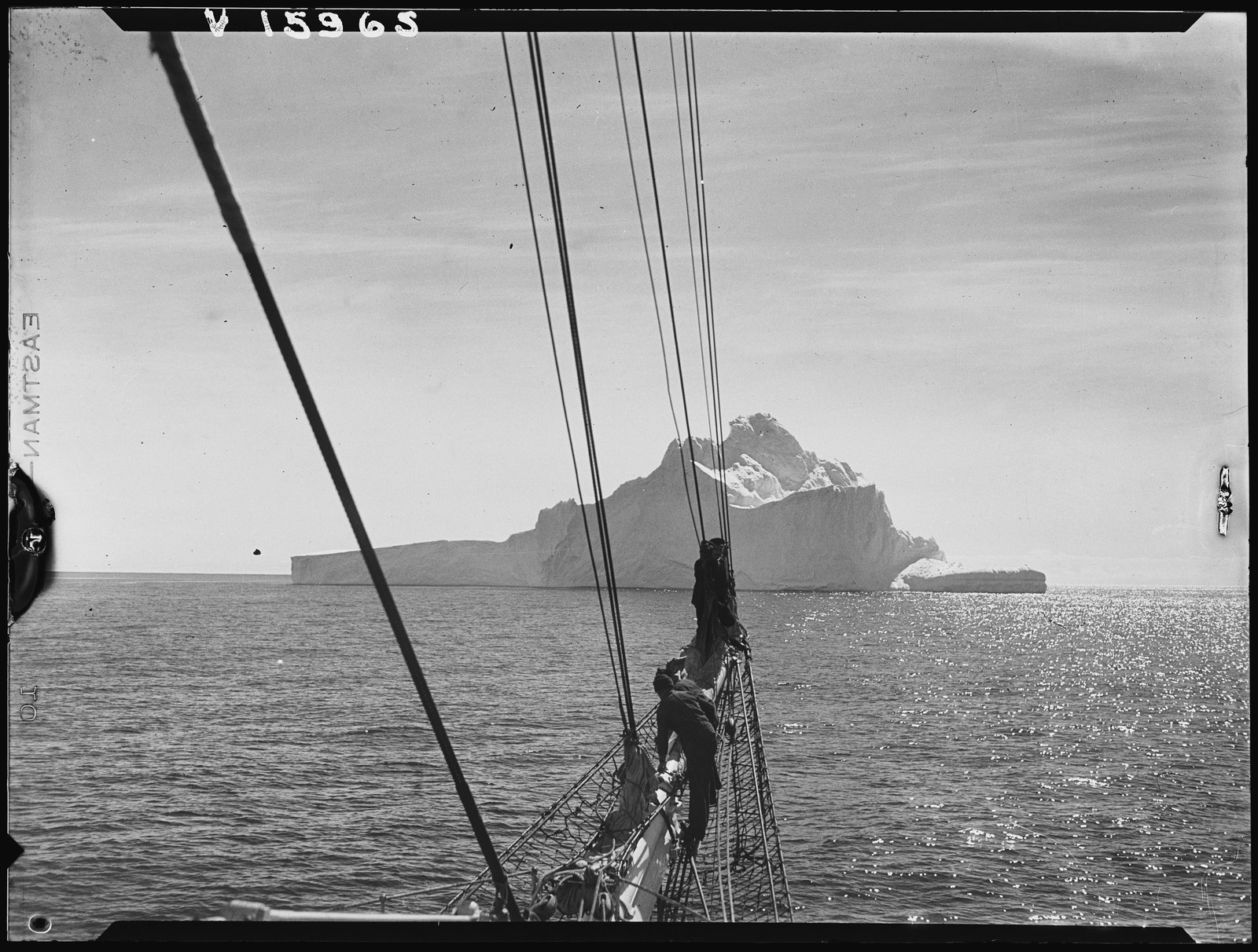 Black and white photograph of men on a ship's bow, facing an iceberg.