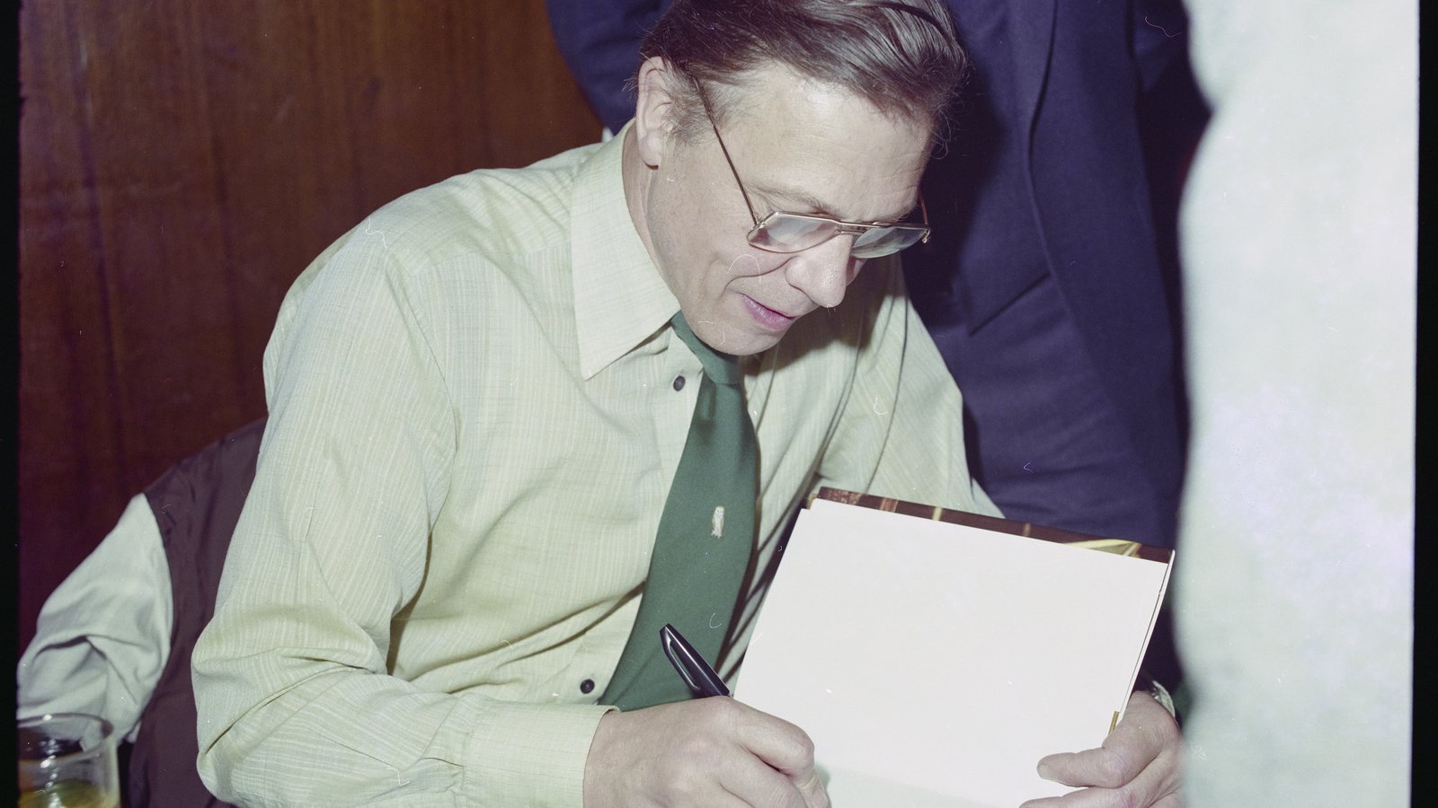 Man in a green shirt and tie signing books at a table, with people around him.