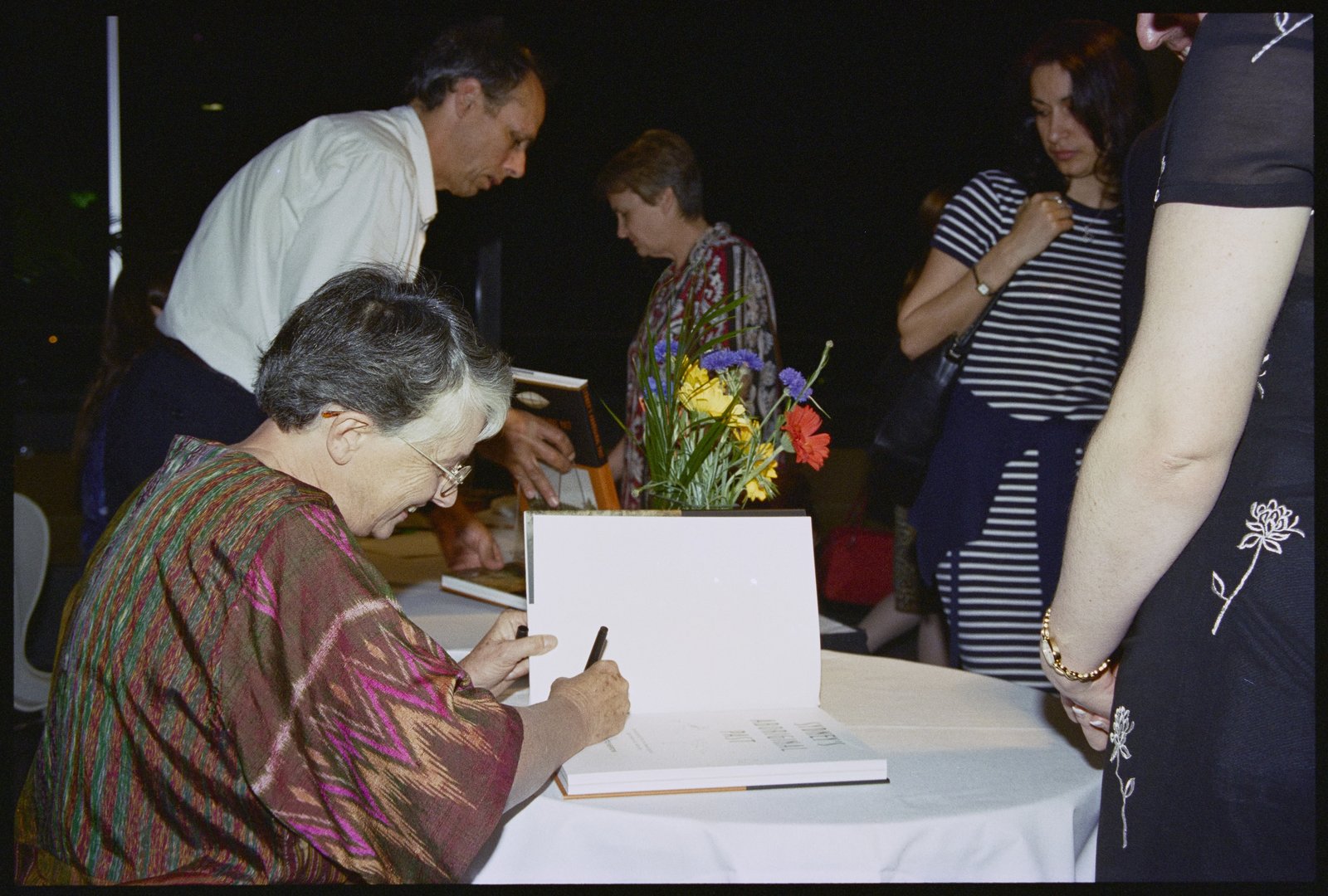 Photograph of a smiling woman in a colourful dress signing a book, with a crowd around her.