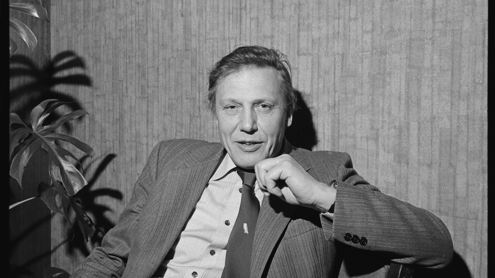 Black and white photograph of a man sitting in a chair, with timber background.