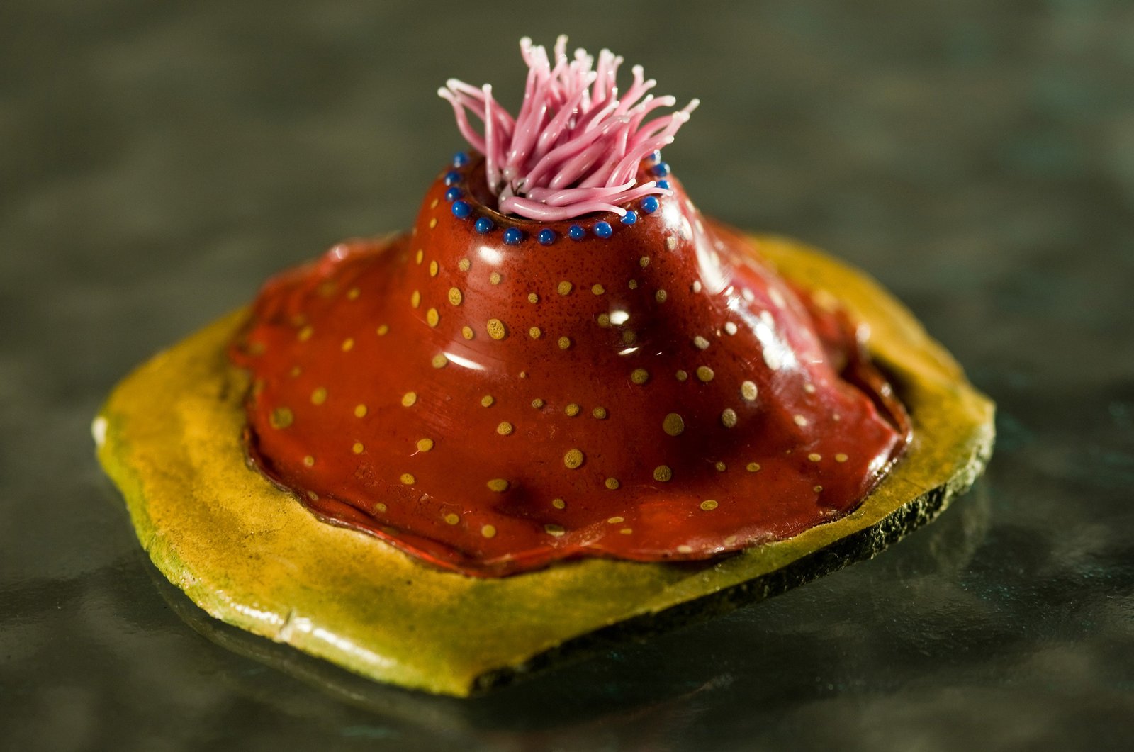 A detailed glass model of a sea anemone with a red cone-shaped body, gold spots and pink tentacles.