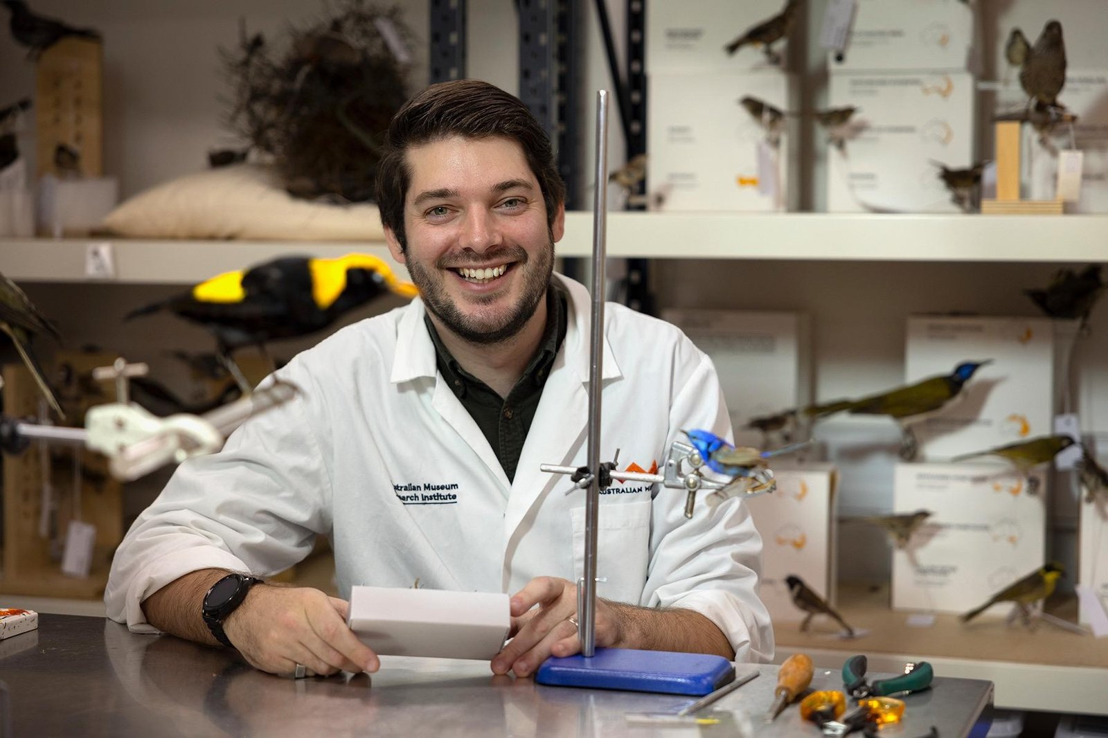 Alex Romano wearing a lab coat whilst sitting at a desk, with bird specimens behind him.