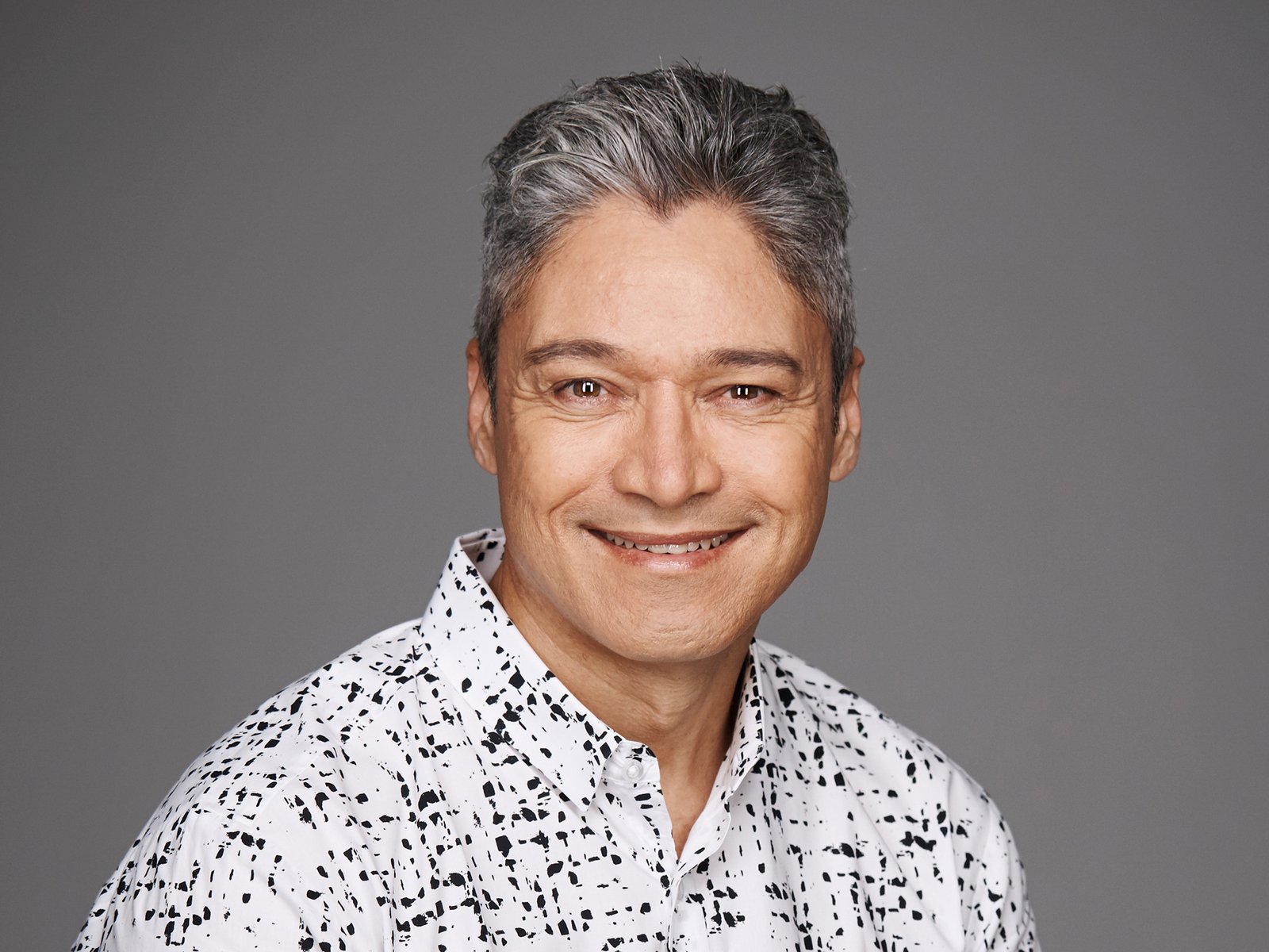 A man with short grey hair smiles at the camera wearing a white patterned shirt.
