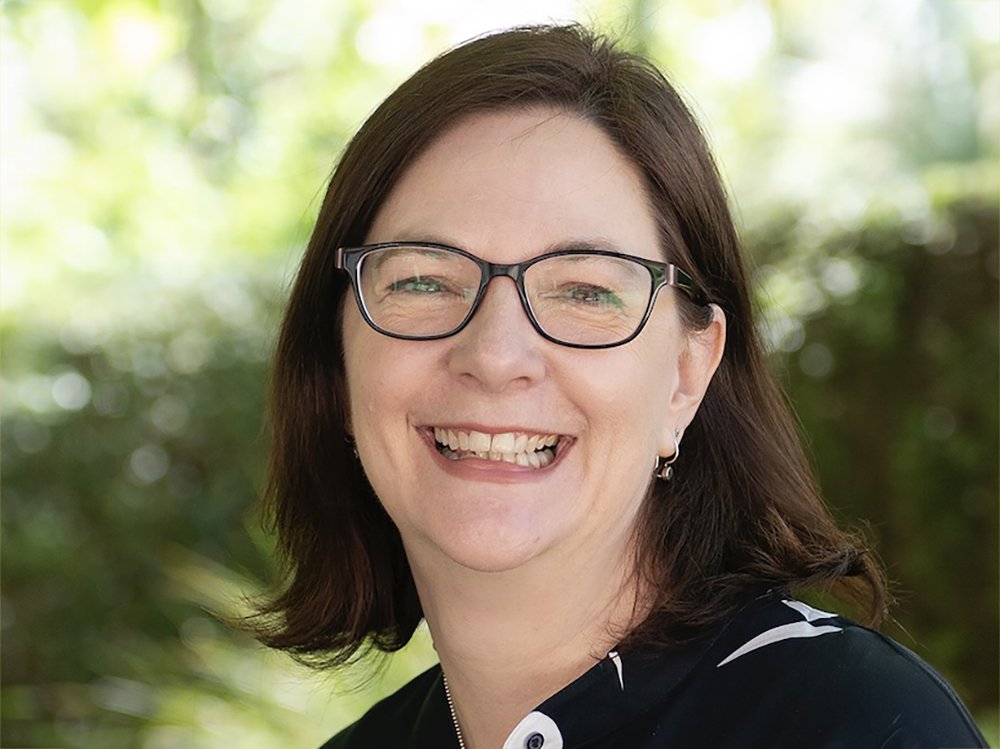 Professor Ariadne Vromen smiling, wearing glasses, photographed outdoors against green foliage