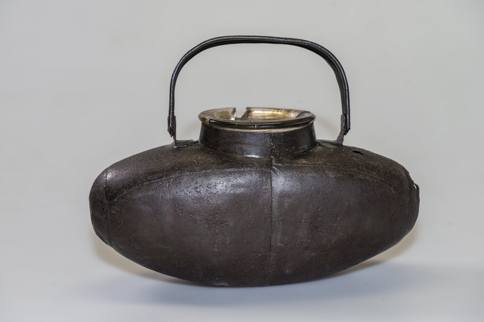 A flattened oval glass bottle in a dark iron protective case with a metal handle against a grey back