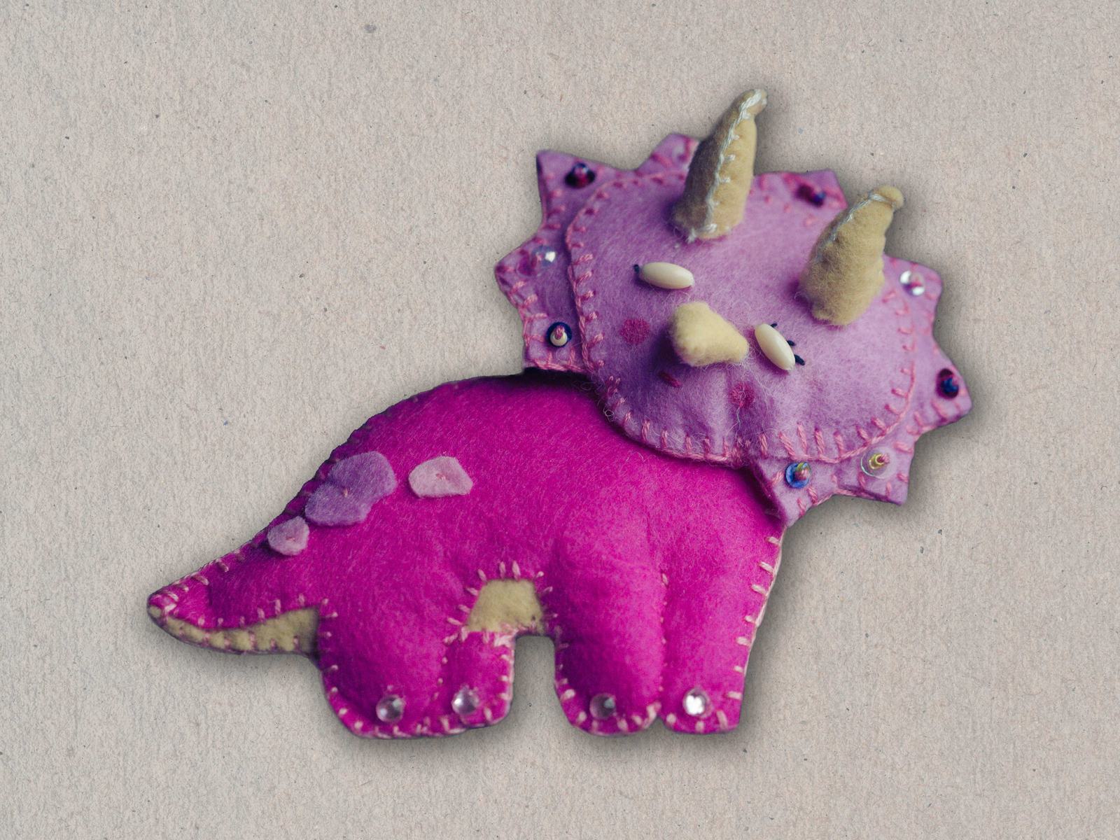 Felted craft of a pink triceratops toy viewed from above, showing two large horns, a frill, and decorative studded spikes along its back and legs.