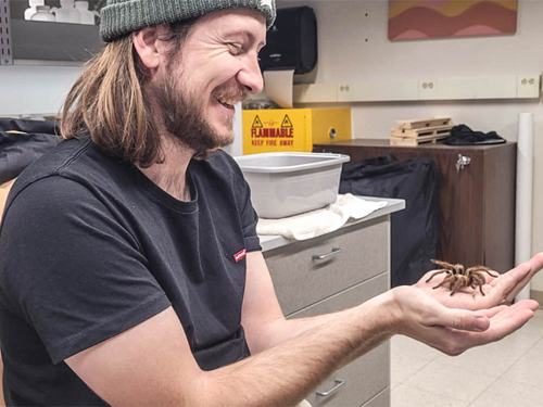 Scientist with a beanie and a beard is holding a tarantula in a lab.