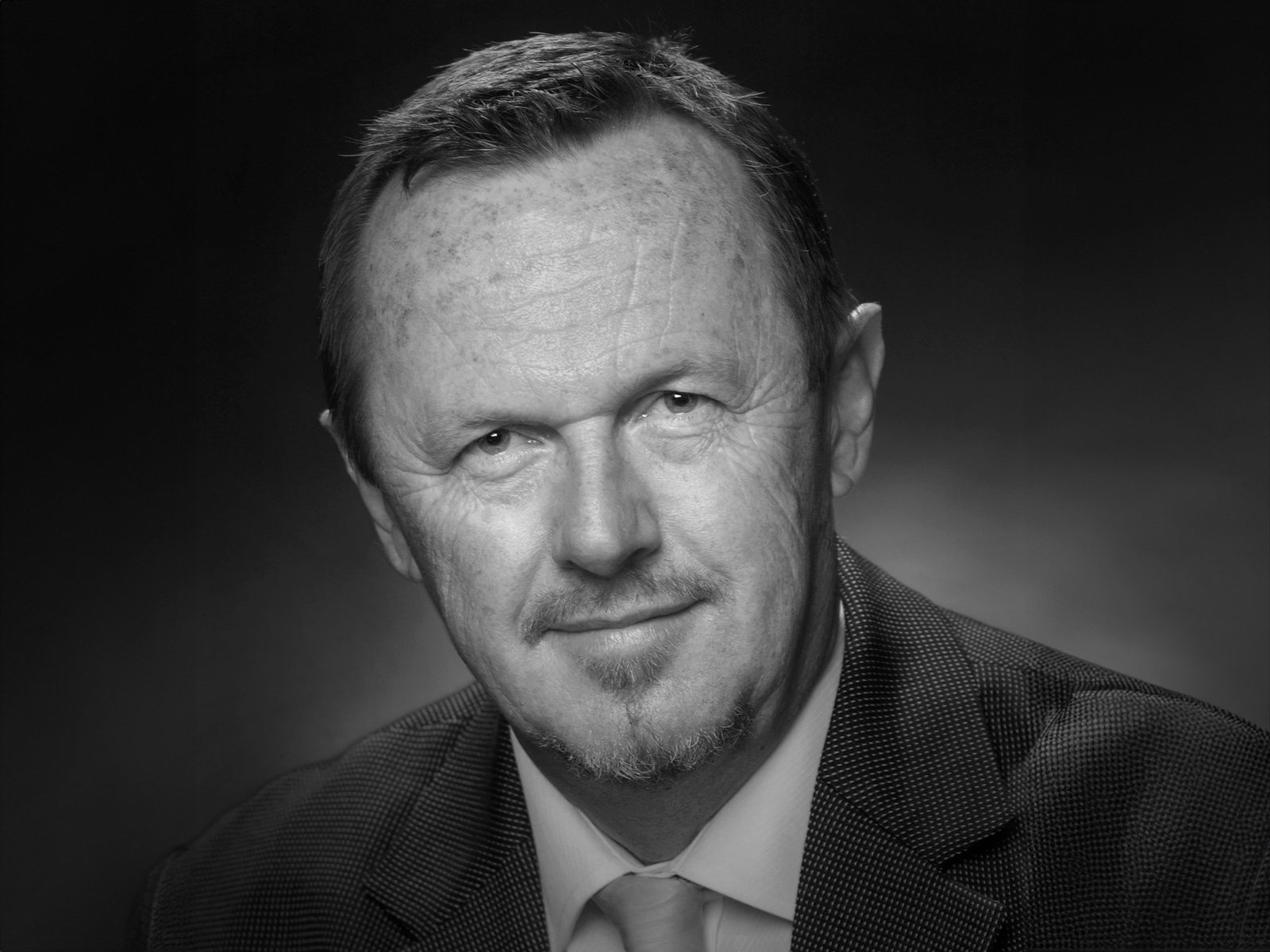 A black and white portrait of a man with short hair wearing a suit jacket against a dark background.