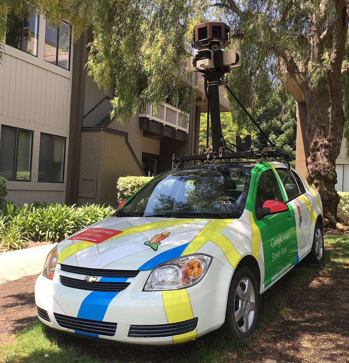 A car with a large black rod fixed to its roof with a camera attached, buildings and trees in view
