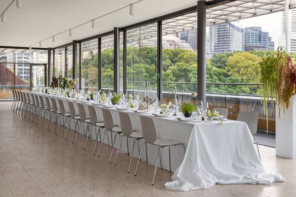Long white-clothed banquet table set with florals along a glass wall overlooking city skyline and trees.
