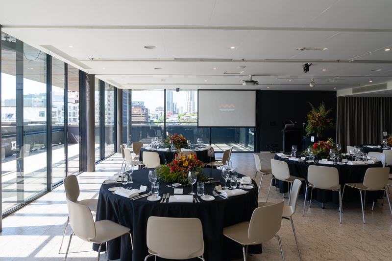 Conference room with large windows, tables and chairs arranged in a U shape, a projector screen, and flower arrangements on the tables.