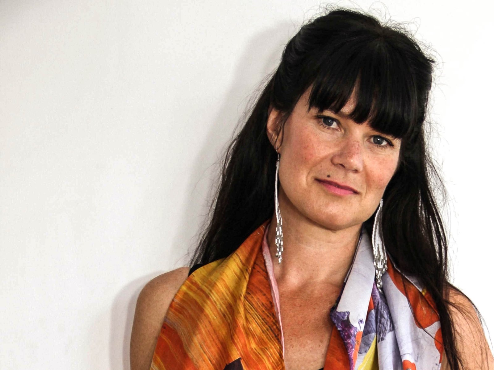 A woman with dark hair and a fringe smiles at the camera wearing a colourful top and long earrings.