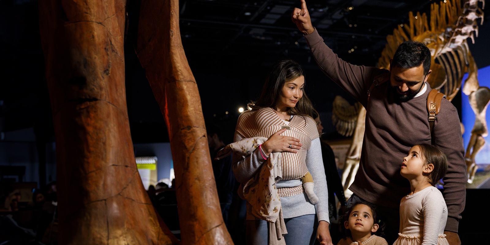 Family looks up in awe at towering dinosaur skeleton legs, father pointing upward beside height measurement chart in exhibition.
