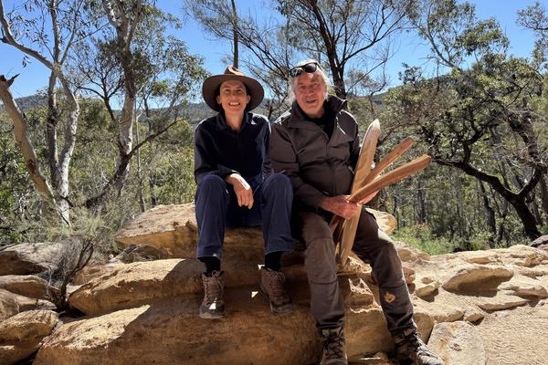 Two people seated on sandstone rocks in bushland in the Blue Mountains, holding carved wooden objects.