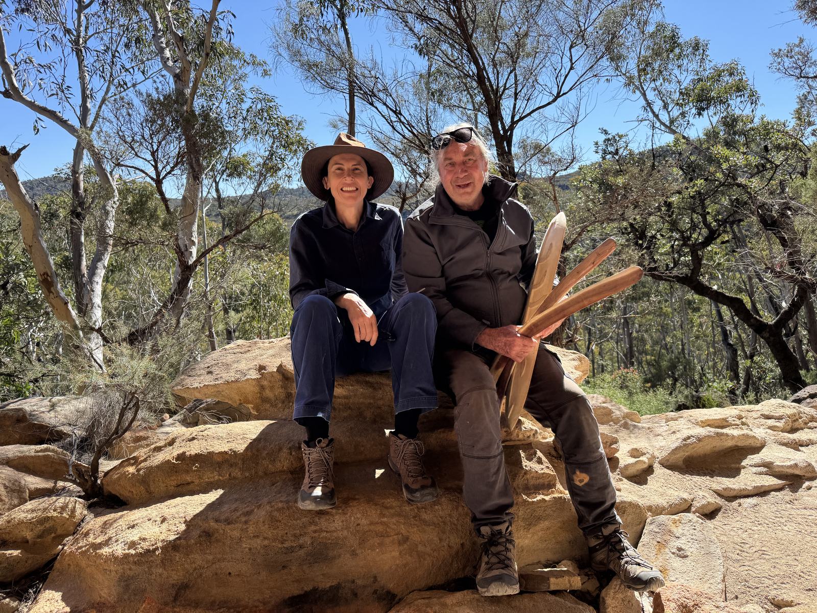 Two people seated on sandstone rocks in bushland in the Blue Mountains, holding carved wooden objects.