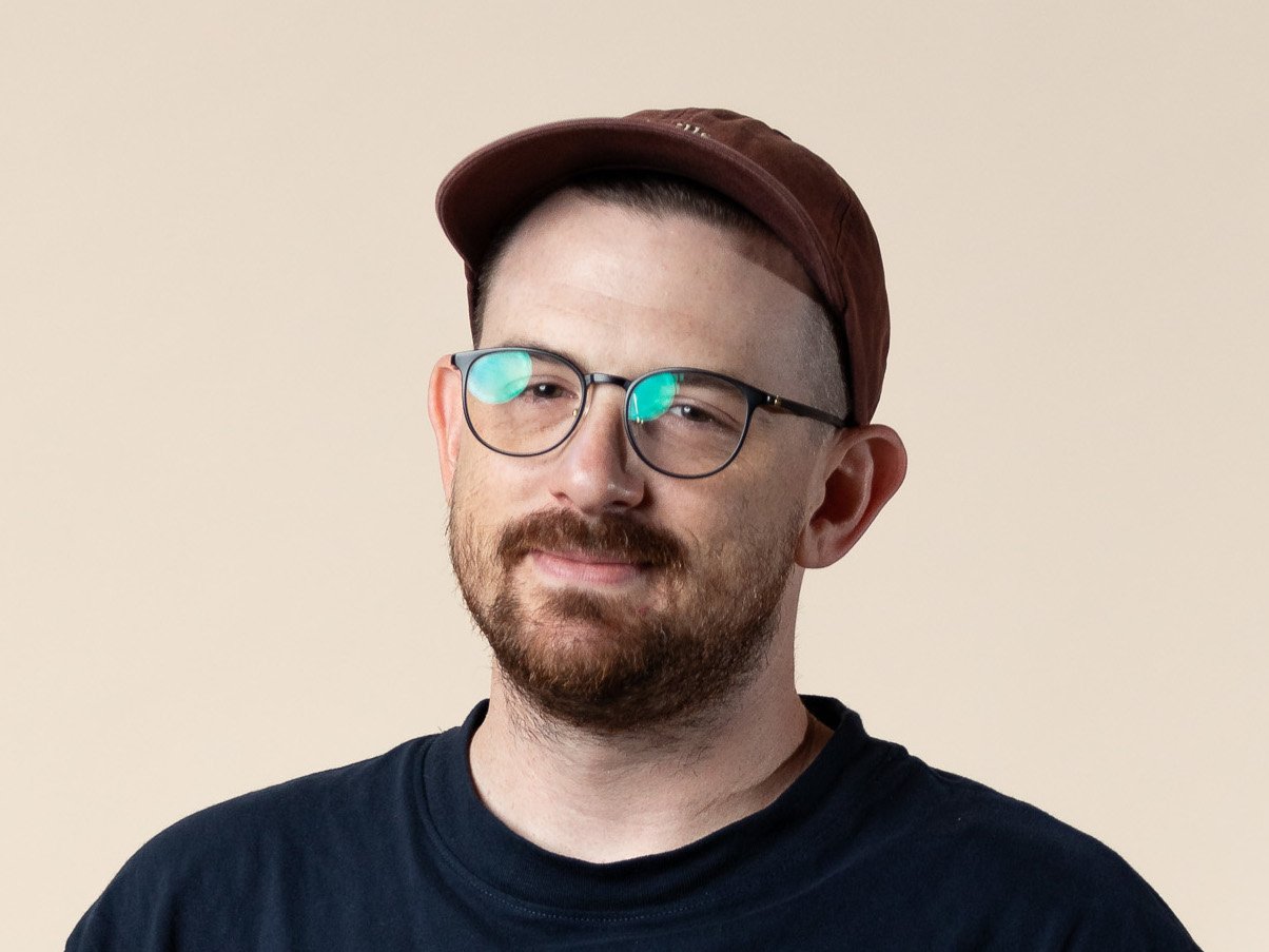 A person with a beard and glasses wearing a dark t-shirt and brown cap against a beige background.