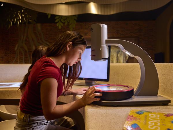 A woman in a red shirt sits at a desk using a microscope to examine something on a screen.