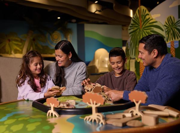 Family of four engaged in an interactive exhibit, smiling and looking at a model of a forest with wooden trees and animals on a table.