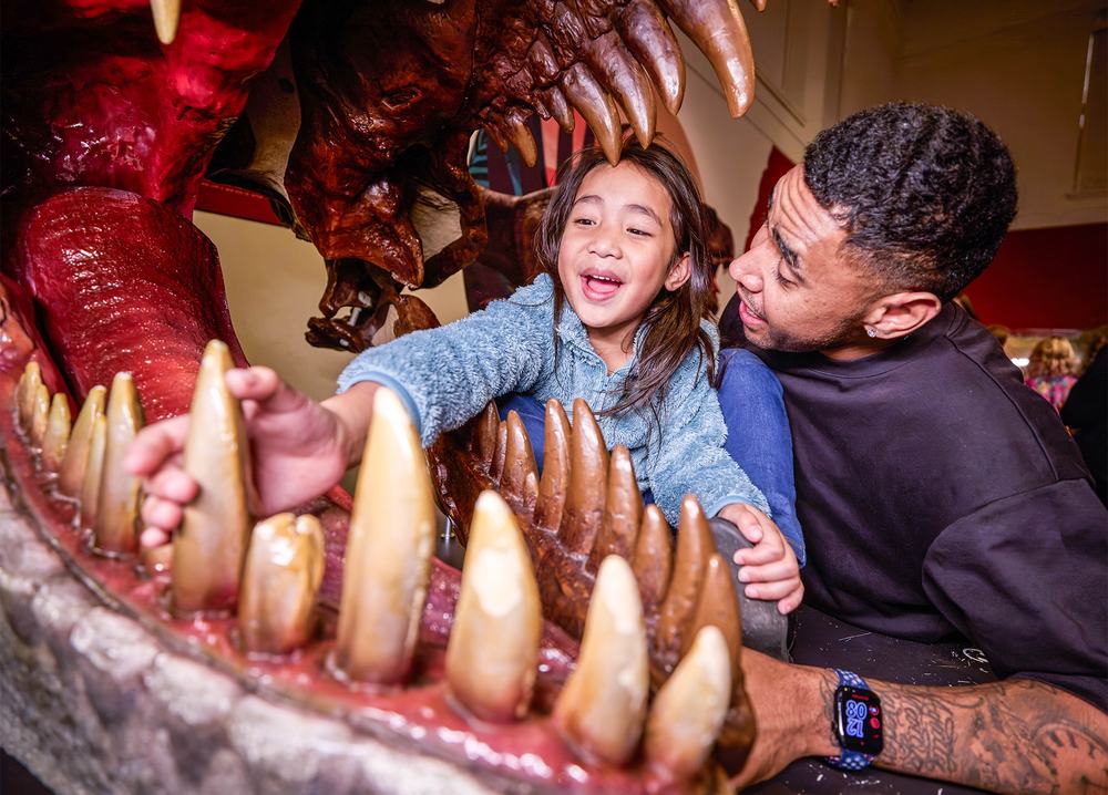 Visitor and child interact with a large dinosaur model, framed by open jaws and teeth inside the Australian Museum.