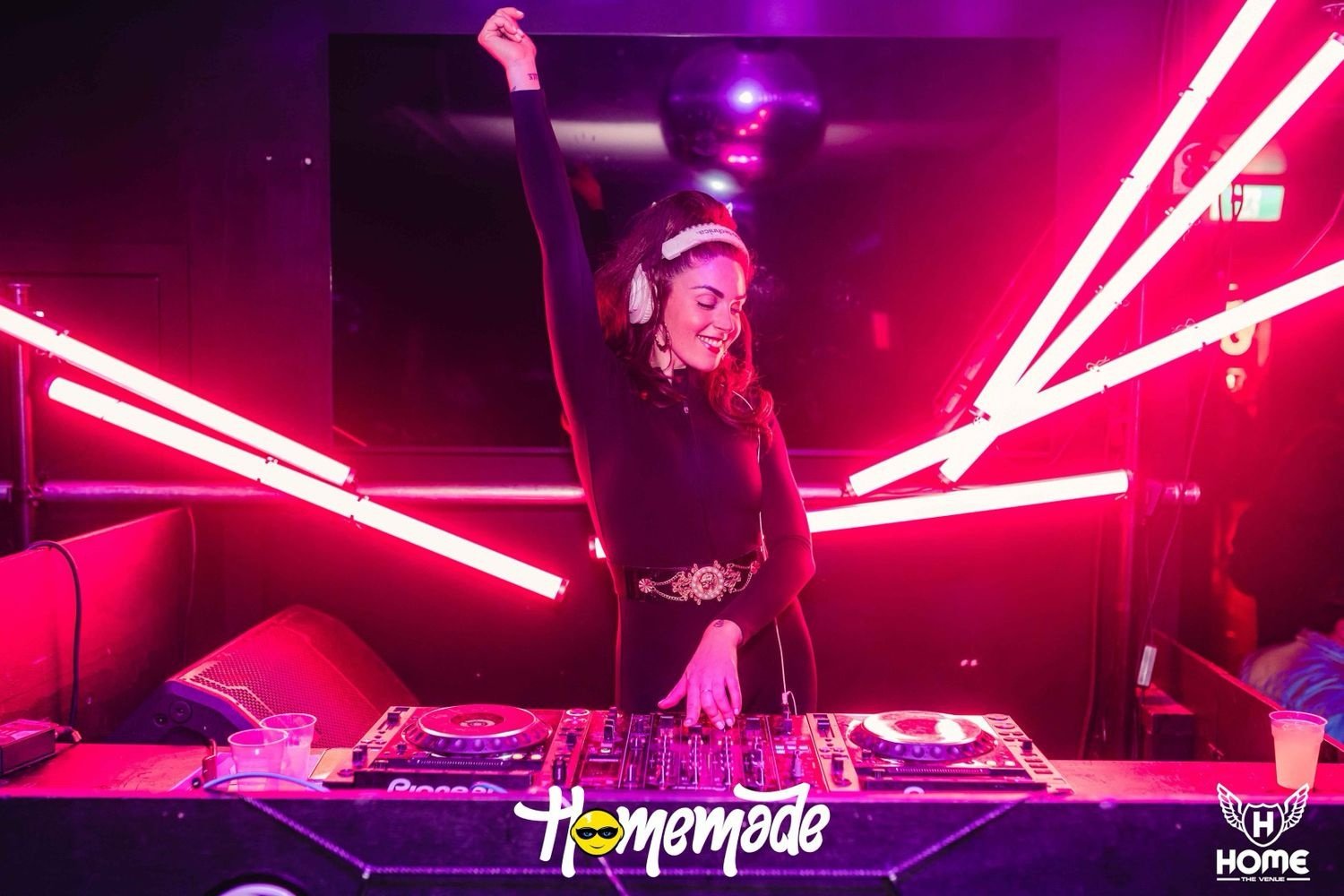 A DJ raises one arm above a Pioneer decks setup under pink neon lights at the Homemade club night.
