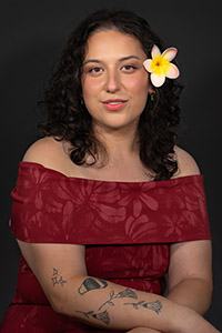 Woman with curly hair and a frangipani flower smiles, wearing a red floral off-shoulder dress.