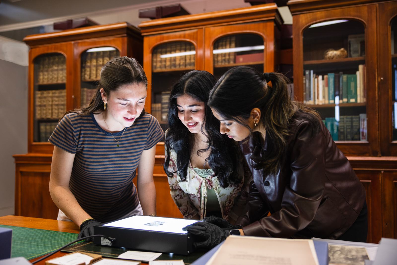 Three students standing around a lightbox, with a photographic negative. Behind them are old bookshelves.