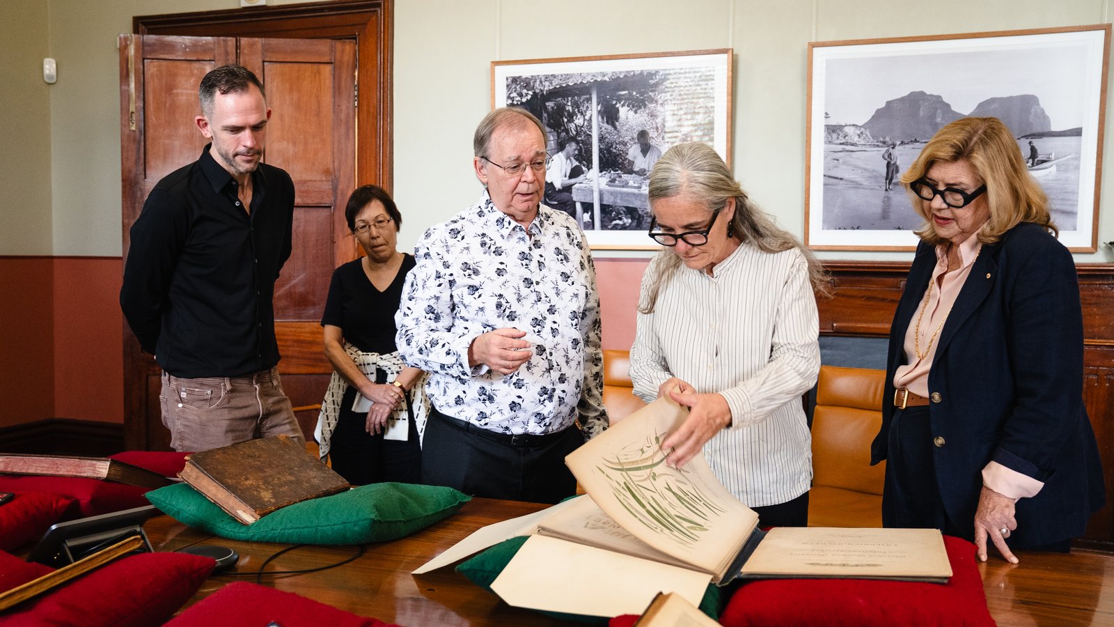 Five people standing around a table with old books on pillows. Artworks in background.