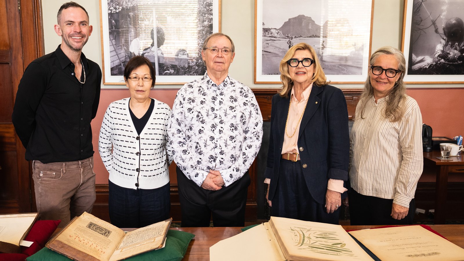 Five people standing around a table with old books on pillows. Artworks in background.