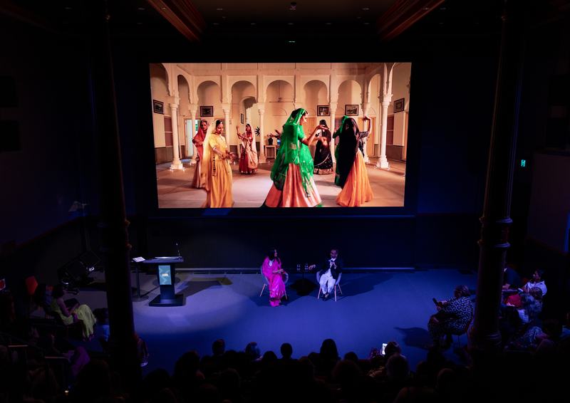A large screen showing a scene with several people in traditional Indian dress with speakers on stage addressing a seated audience.
