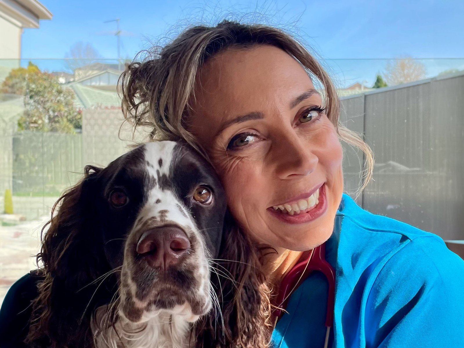 A smiling woman in a blue veterinary uniform poses cheek-to-cheek with a spaniel outdoors.
