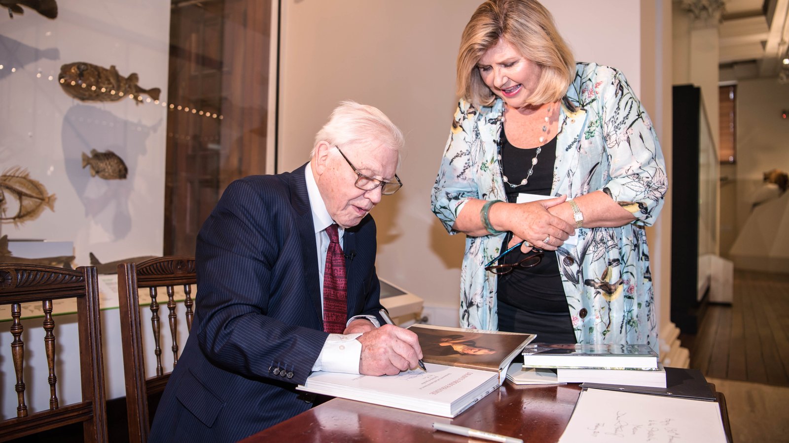 Sir David Attenborough signs a book at a table next to Australian Museum Director & CEO Kim McKay