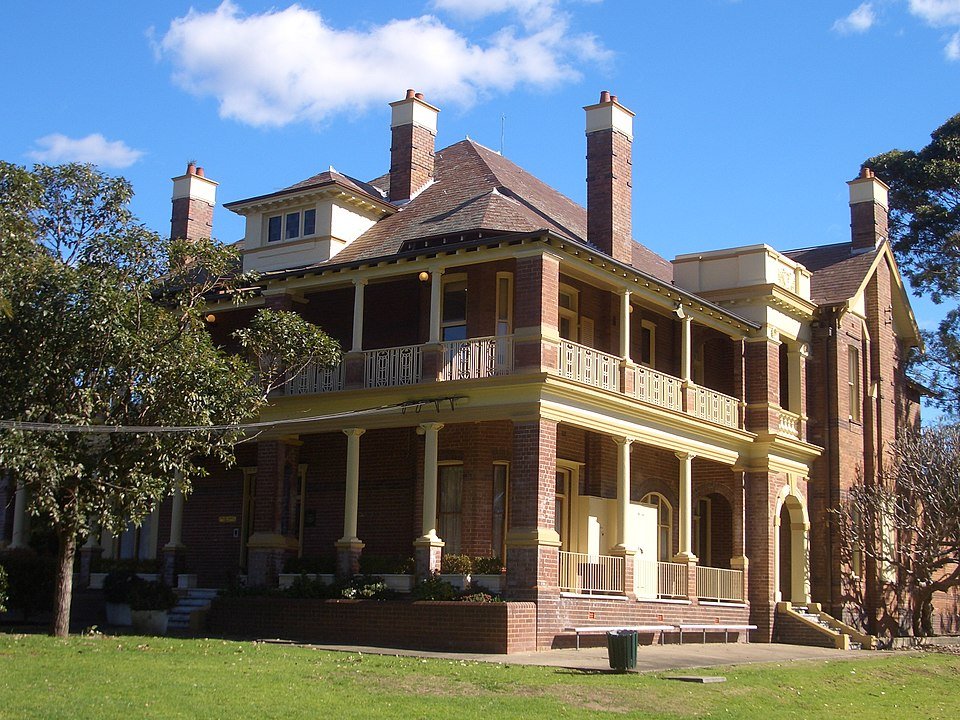Two‑storey historic house with arched verandas and a tiled roof, partially obscured by trees.