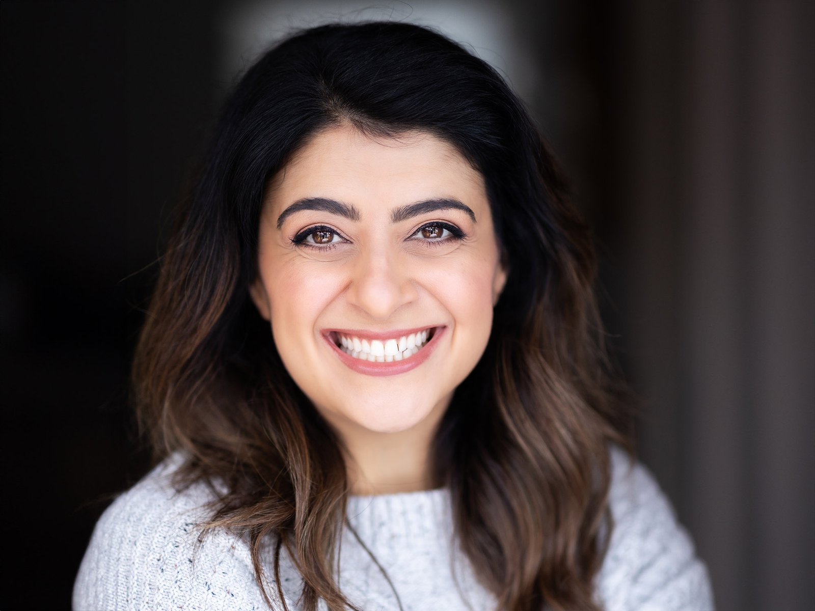 A woman with long dark hair smiles broadly at the camera wearing a light grey top.