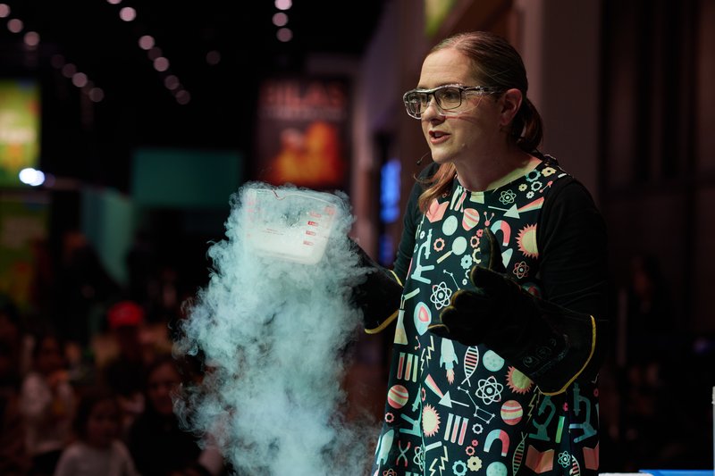 A science presenter holds a smoking container while demonstrating an experiment to an audience