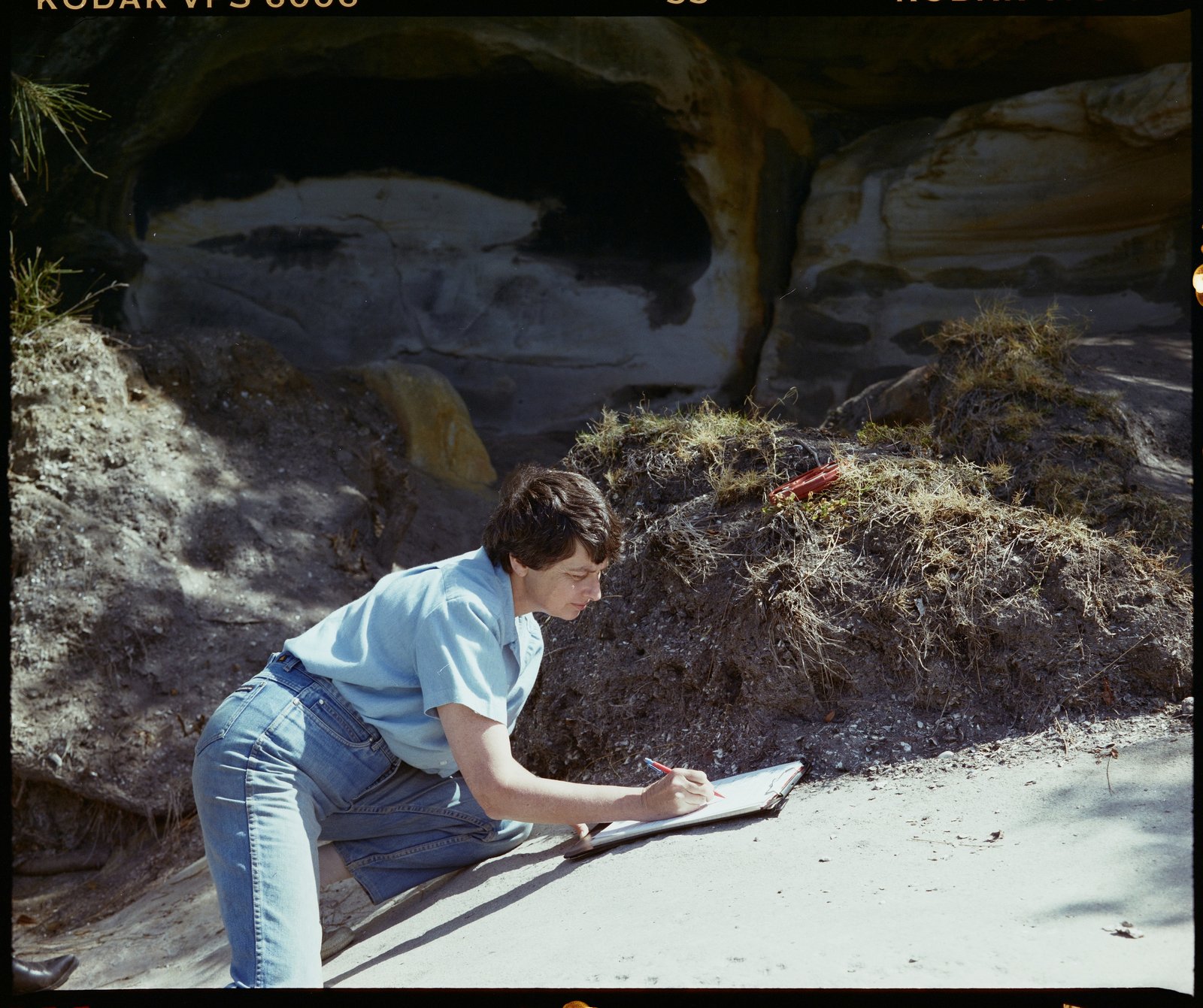 Woman in jeans writing on a notepad, on a rock. A cave is in the background.