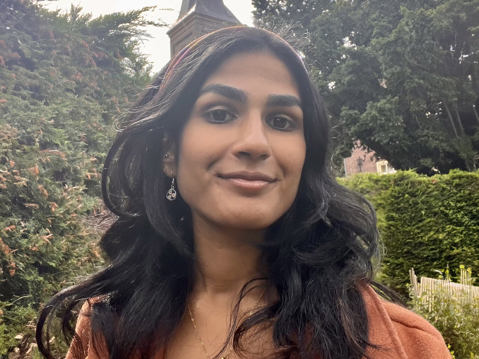 A woman with long dark hair and drop earrings smiles at the camera wearing an orange top