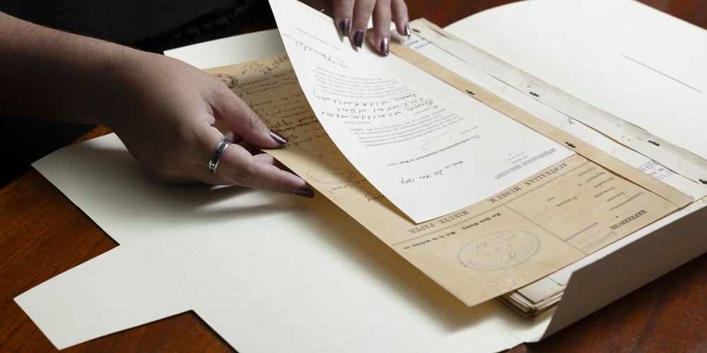 A person lifts a handwritten letter from an open folder of documents from the museum archives