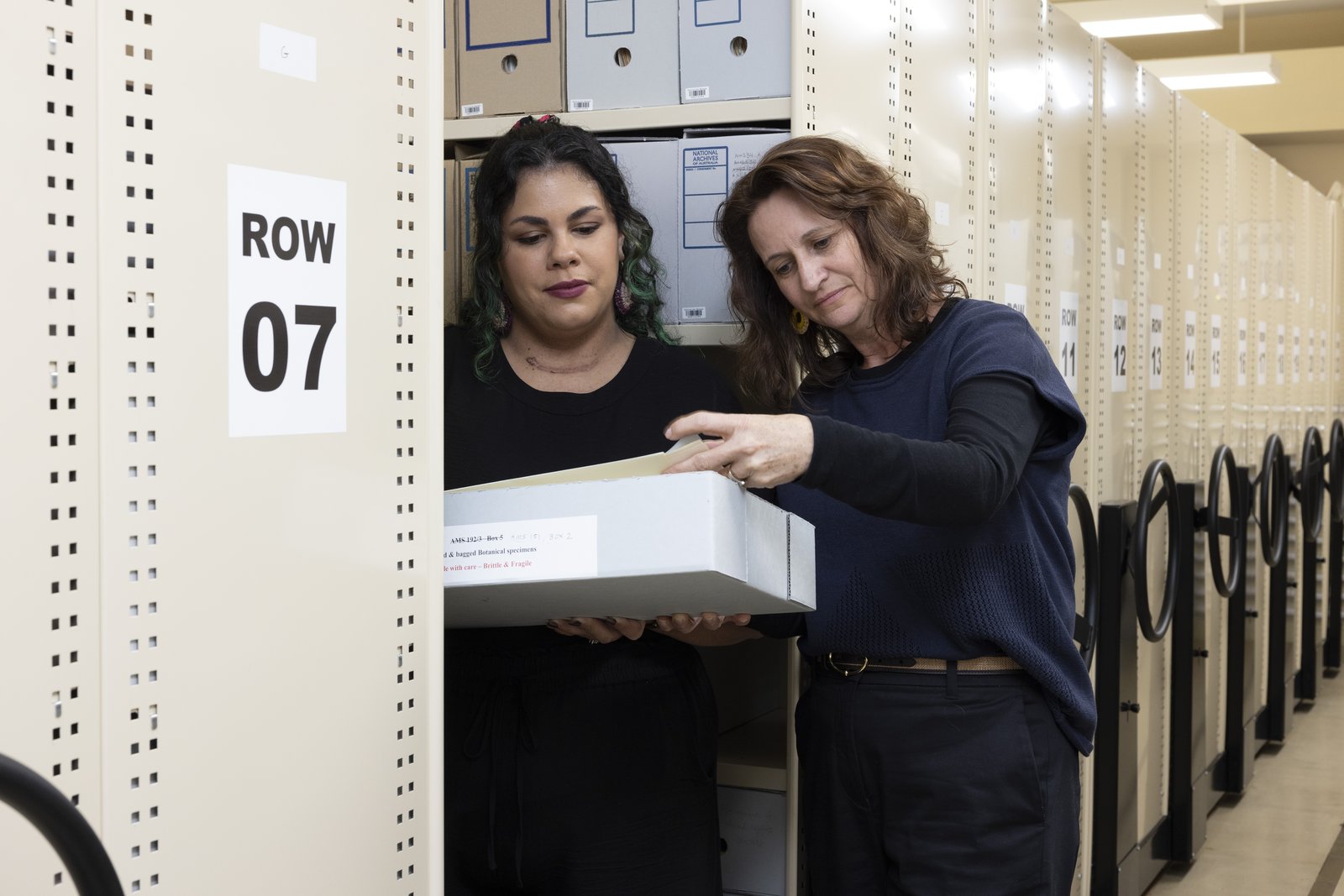Two women examine a labelled archival box in large collection area with numbered rows.