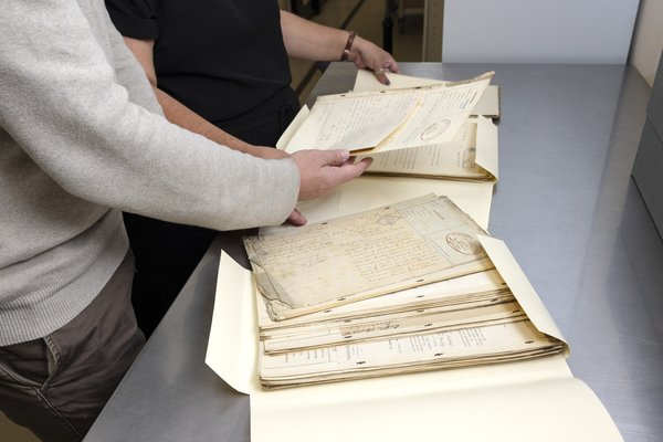Two people handle a stack of aged handwritten documents and letters in an archival setting.
