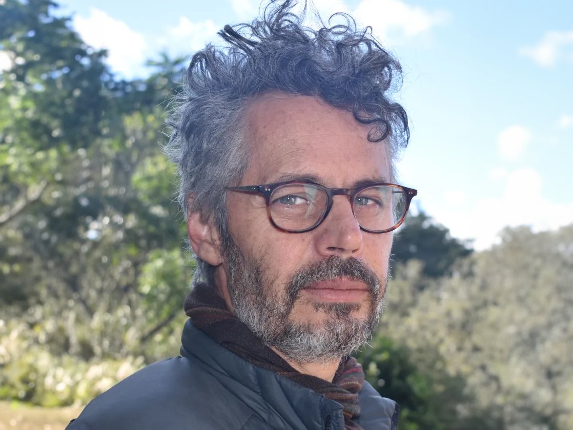 A man with curly grey hair and a beard wearing glasses and a grey jacket outdoors.
