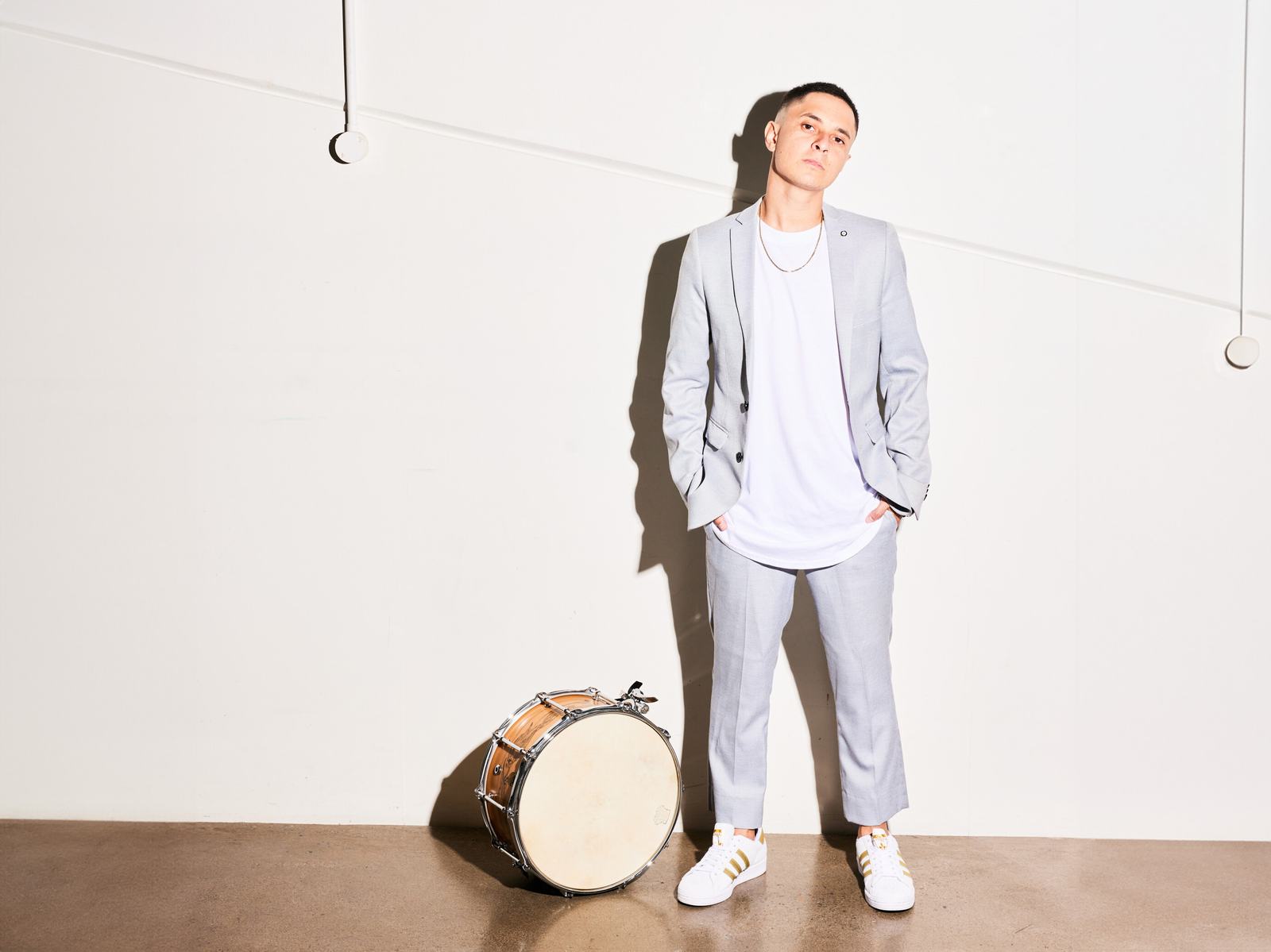 Man in a grey suit stands beside a drum, hands in pockets, against a white wall.