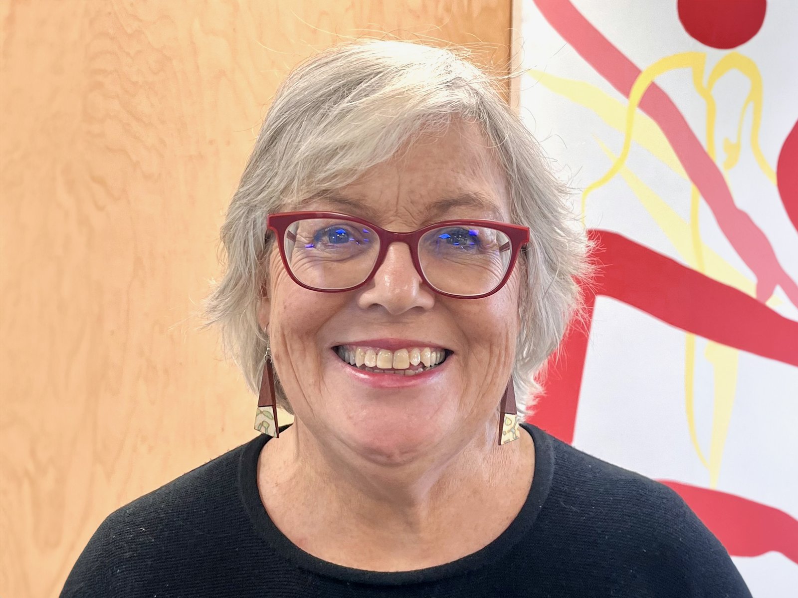Professor Bronwyn Hayward smiling, wearing red-framed glasses, against a decorative wall mural